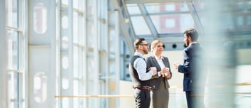 Three business professionals in formal attire are talking and holding cups in a bright, modern office space with large windows.