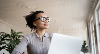 Woman with glasses sitting at a table, looking thoughtful, with an open laptop in front of her. Indoor setting with plants and ornate ceiling in the background.