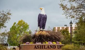A large eagle statue painted purple and white stands on a brick base with "ASHLAND UN" visible, surrounded by plants and flowers on a university campus.